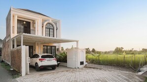 A two-story house with a covered driveway, a parked car, and a fenced field in the background. - Been View Homestay (Berbah)