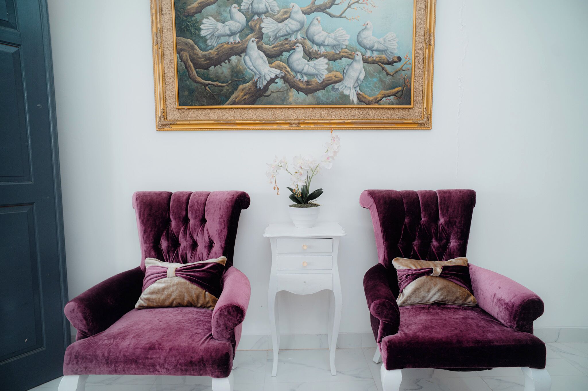 Dos sillones morados y mullidos con cojines decorativos, una mesita de noche blanca con una planta, y un cuadro con pájaros en un árbol en la parte superior.