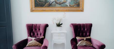 Dos sillones morados y mullidos con cojines decorativos, una mesita de noche blanca con una planta, y un cuadro con pájaros en un árbol en la parte superior.