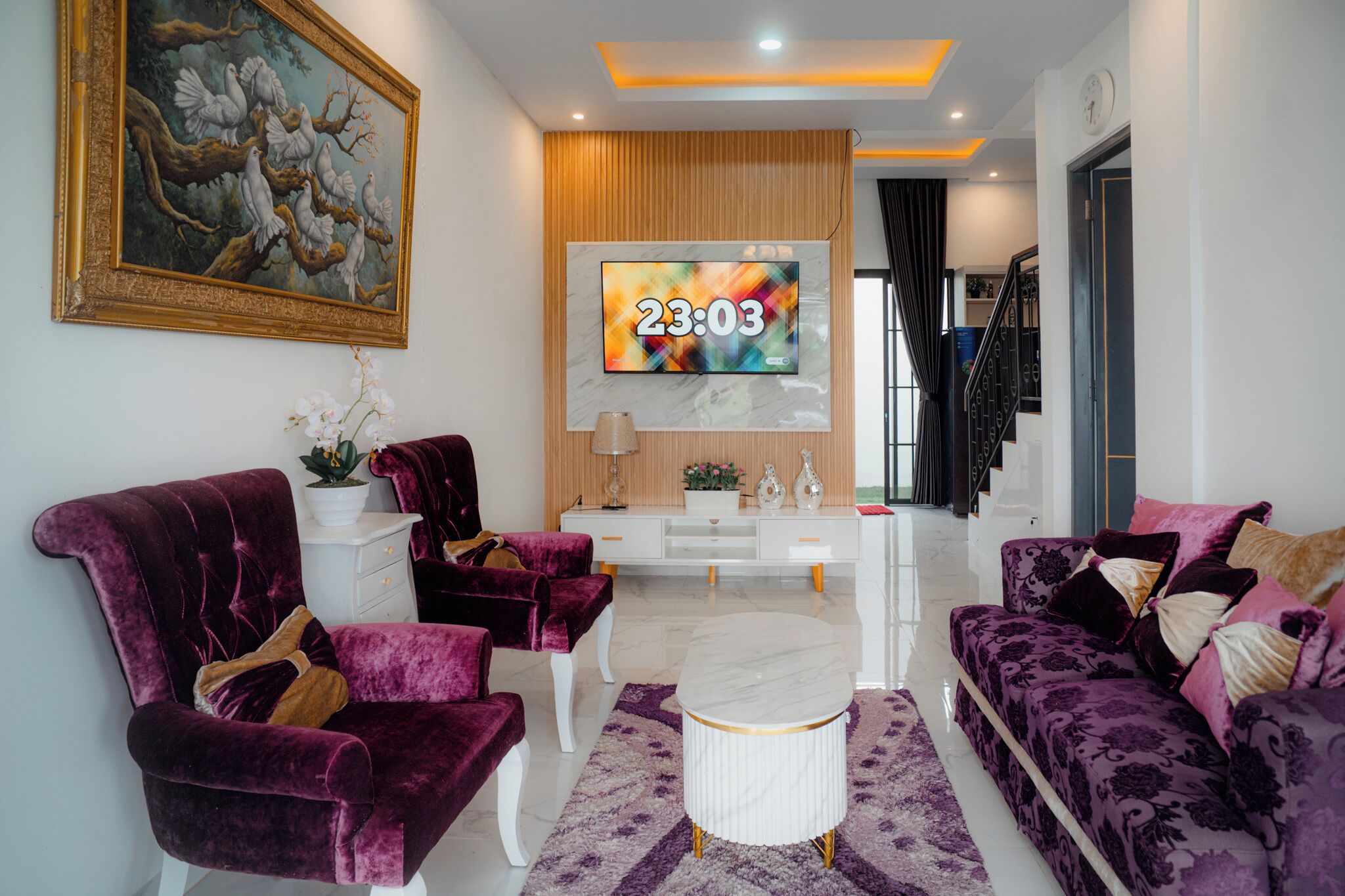 A modern living room with a large TV, two purple sofas, a white coffee table, and a painting of doves on the wall.