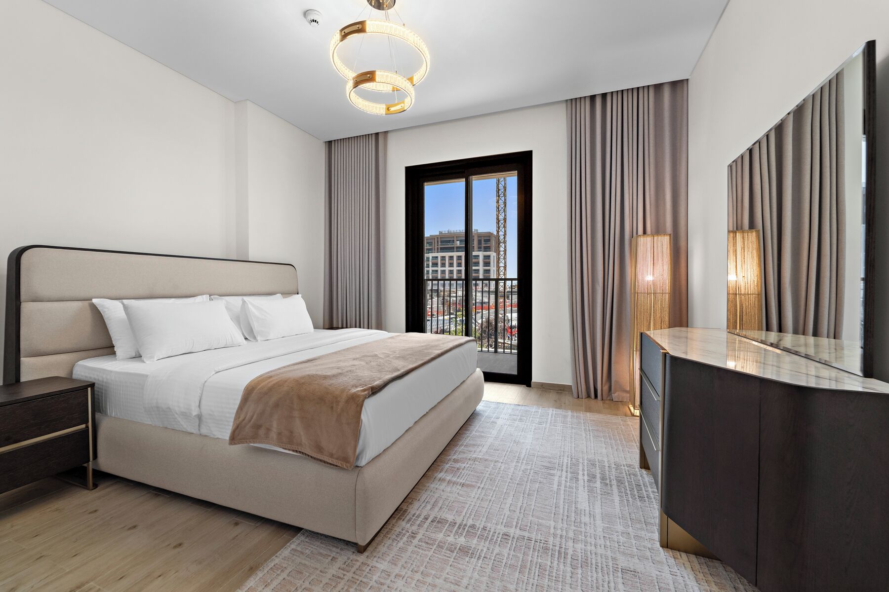 A modern hotel room with a large bed, a bedside table, a vanity with a mirror, and a view of the city through a large window.