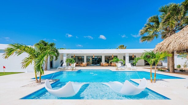 A modern resort with a large swimming pool, palm trees, and a thatched-roof building. - Conch Beach Villa NEW 7-Bed 6 Bath Pool 5-min to Beach by Bocobay (Noord)