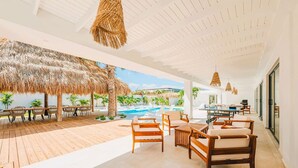 An open-plan living area with a pool, wooden furniture, and a thatched ceiling. - Conch Beach Villa NEW 7-Bed 6 Bath Pool 5-min to Beach by Bocobay (Noord)