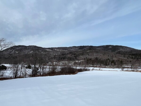 A snowy landscape with a flat, open field in the foreground and a forested hill in the background. - Mountain Retreat With River Views Near Sunday River and Year-round Adventure (Rumford)