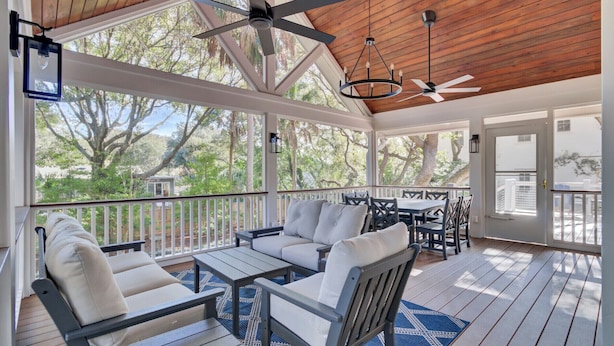 A covered outdoor seating area with a wooden ceiling, white furniture, and a view of trees and a house in the background. - Steps to Isle of Palms Beach w/ Porch Dining, Outdoor Shower & Open Living (Isle Of Palms)