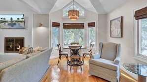 A spacious living room with a fireplace, a dining area with a chandelier, and a large window with a view of trees. - Steps to Isle of Palms Beach w/ Porch Dining, Outdoor Shower & Open Living (Isle Of Palms)