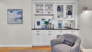 A modern kitchen with white cabinetry, a black countertop, and a built-in island. There is a framed painting on the wall and a chair with a cushion in the foreground. - Steps to Isle of Palms Beach w/ Porch Dining, Outdoor Shower & Open Living (Isle Of Palms)
