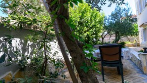 A patio area with a chair, a tree, and a building in the background. - Beachfront apartment fully equipped with outdoor garden space, parking included. (Okrug Gornji)