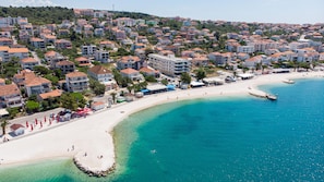 An aerial view of a coastal town with a sandy beach, clear blue waters, and residential buildings. - Beachfront apartment fully equipped with outdoor garden space, parking included. (Okrug Gornji)
