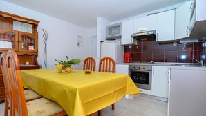 A compact kitchen with white cabinets, a stove, and a microwave. There is a dining table with a yellow tablecloth and wooden chairs. - Beachfront apartment fully equipped with outdoor garden space, parking included. (Okrug Gornji)