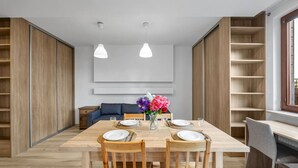 Ein moderner Essbereich mit einem Holztisch, gedeckt für vier Personen, flankiert von Holzstühlen. Im Hintergrund befindet sich eine Couch und ein kleiner Beistelltisch mit einem Blumenstrauß.