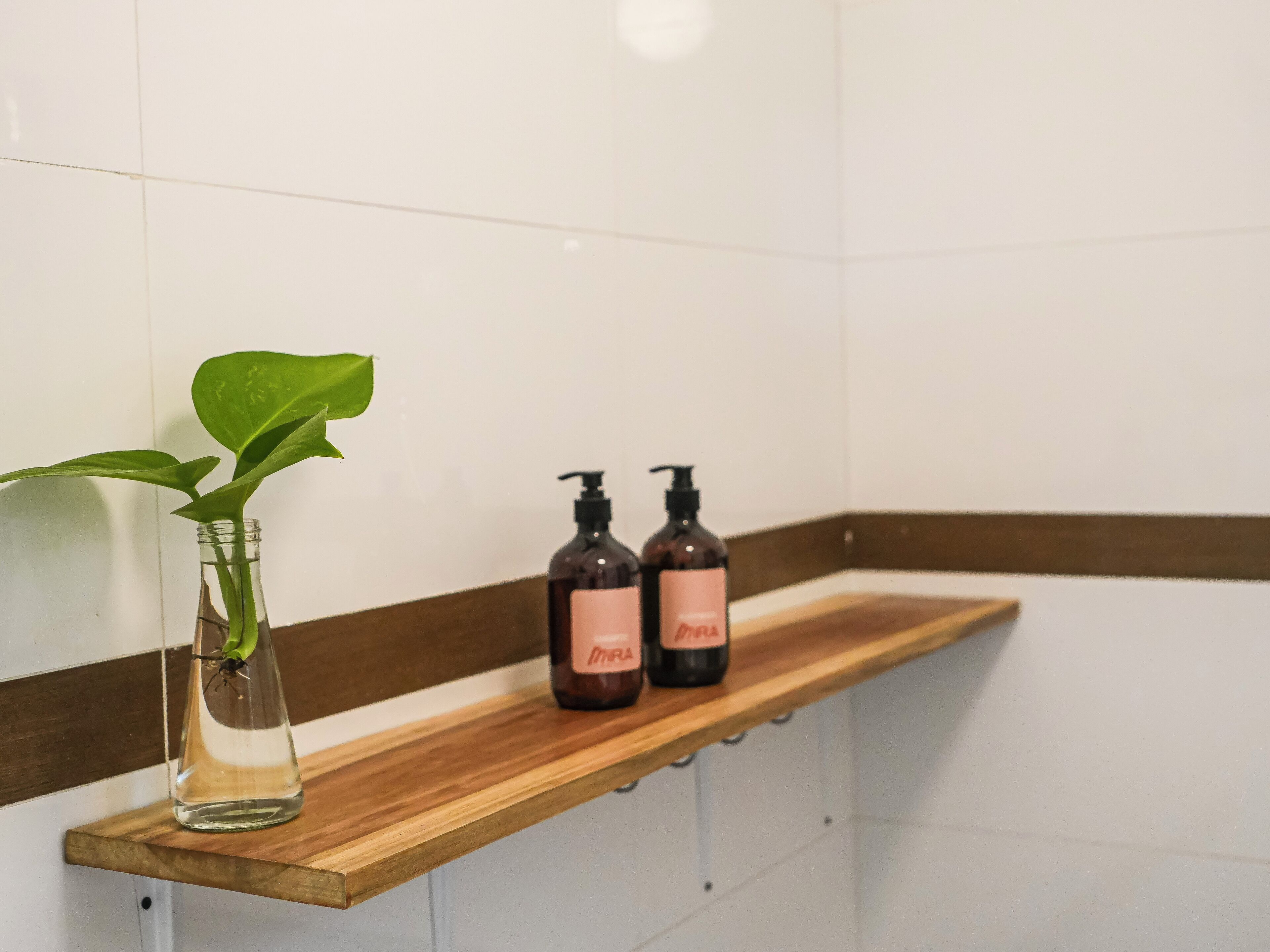 A bathroom with a wooden shelf holding a clear vase with a green plant and two dark bottles with pink labels.
