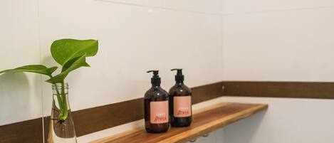 A bathroom with a wooden shelf holding a clear vase with a green plant and two dark bottles with pink labels.