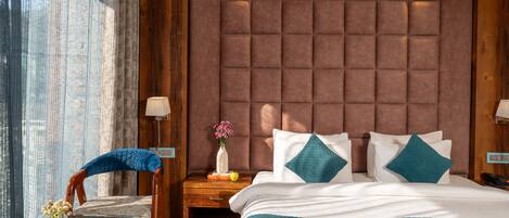 Habitación de hotel con una cama grande, cabecera de madera, una mesa de vidrio con un florero y una lámpara, y una silla con cojín azul.