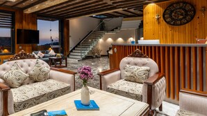 A modern interior with a wooden reception desk, two upholstered sofas with patterned cushions, a marble coffee table with a vase of flowers, and a large wall clock. - Moustache Select Mcleodganj (Dharamshala)