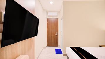 A hotel room with a flat-screen TV mounted on the wall, a wooden door, a white bed with a black and orange bedspread, and a blue rug on the floor.