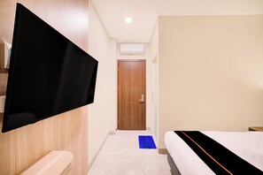 Habitación de hotel con televisor de pantalla plana montado en la pared, puerta de madera, cama blanca con colcha negra y naranja, y alfombra azul en el suelo.