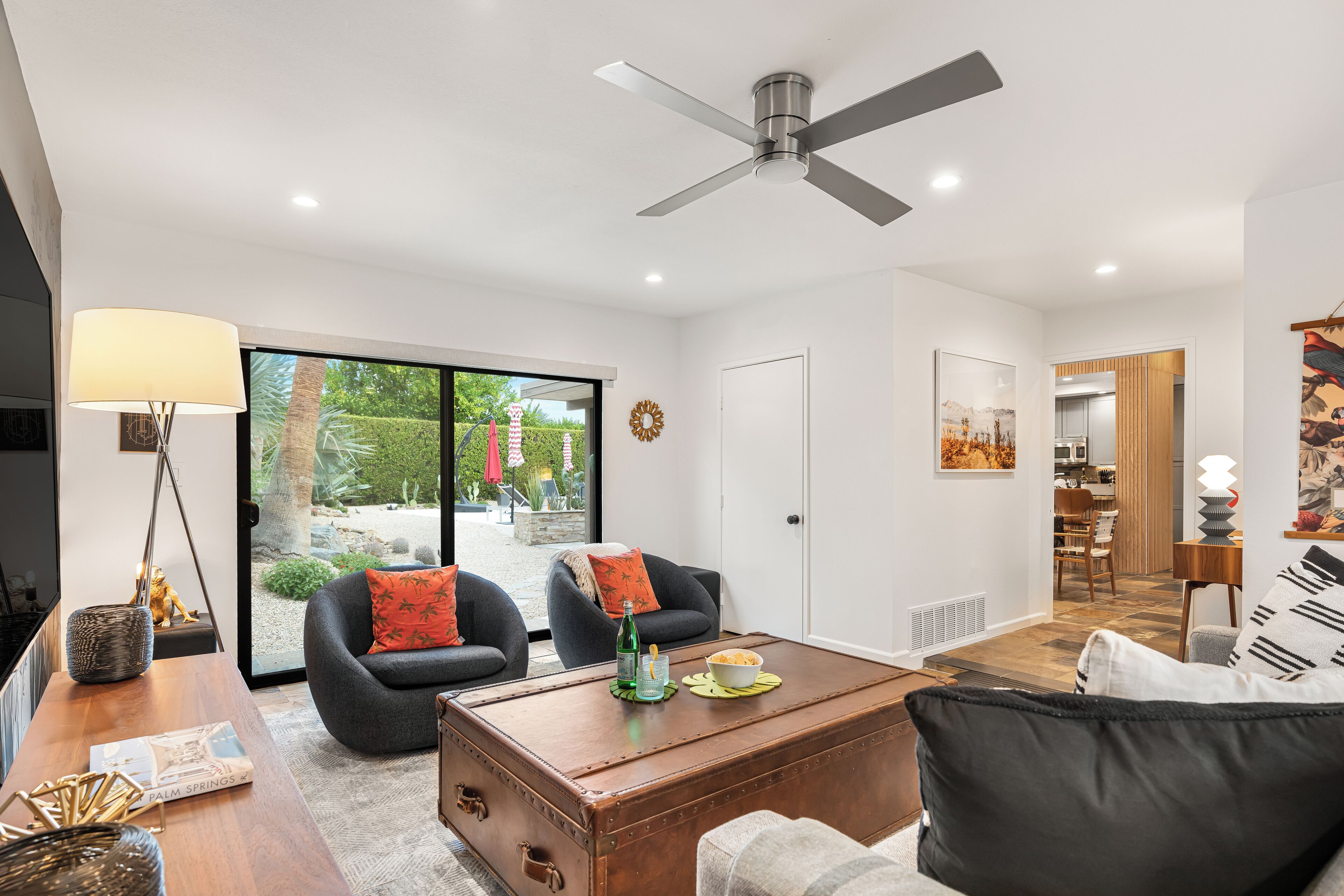 A modern living room with a large window, a ceiling fan, and a dining area visible through the glass doors.