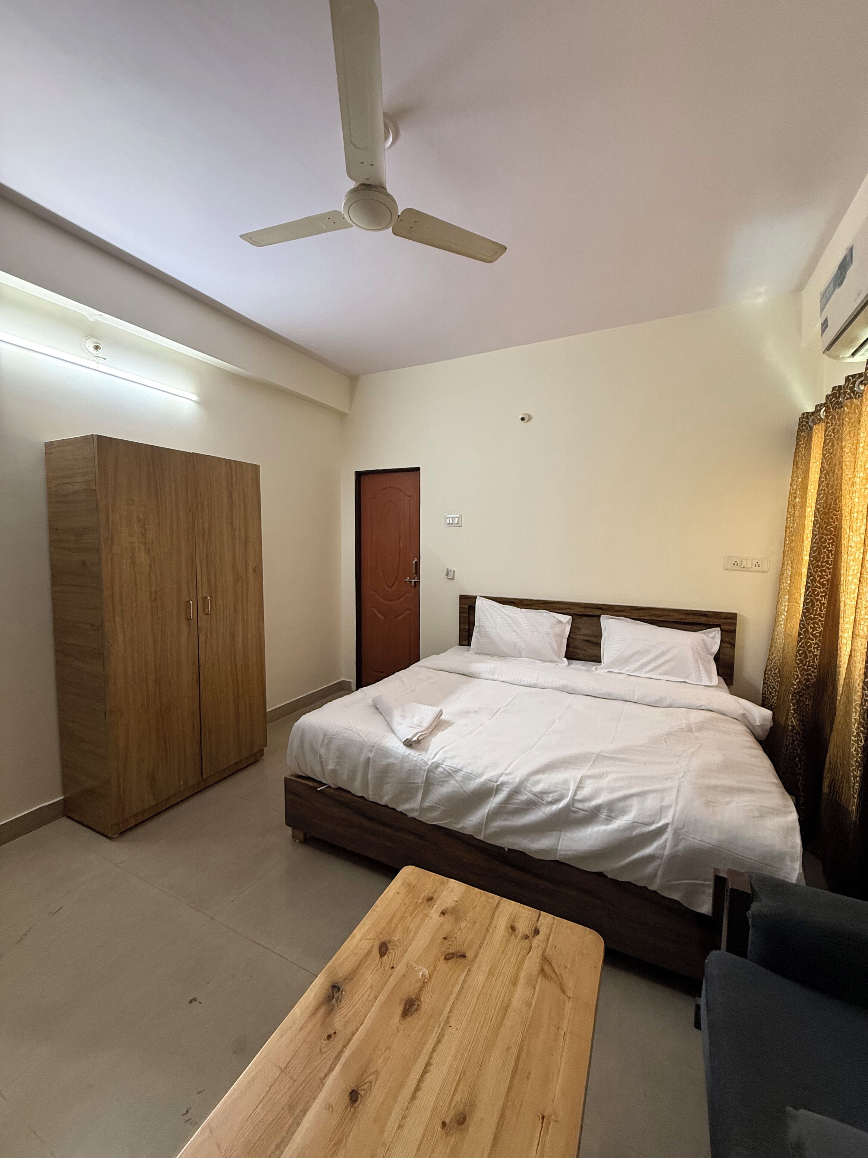 A bedroom with a bed, a wooden wardrobe, a ceiling fan, a wooden coffee table, and a chair.