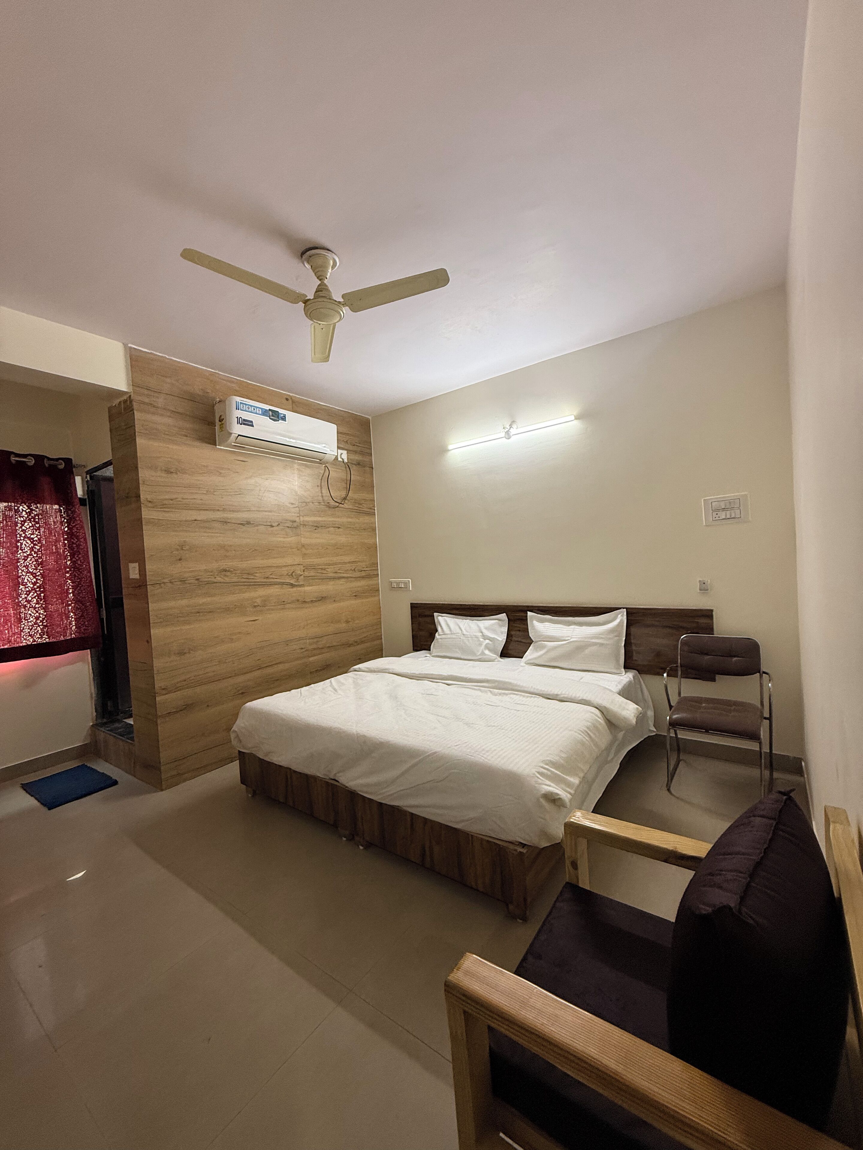 Un dormitorio con cama, ventilador de techo, unidad de aire acondicionado, mesita de noche, una silla y ventana con cortinas.