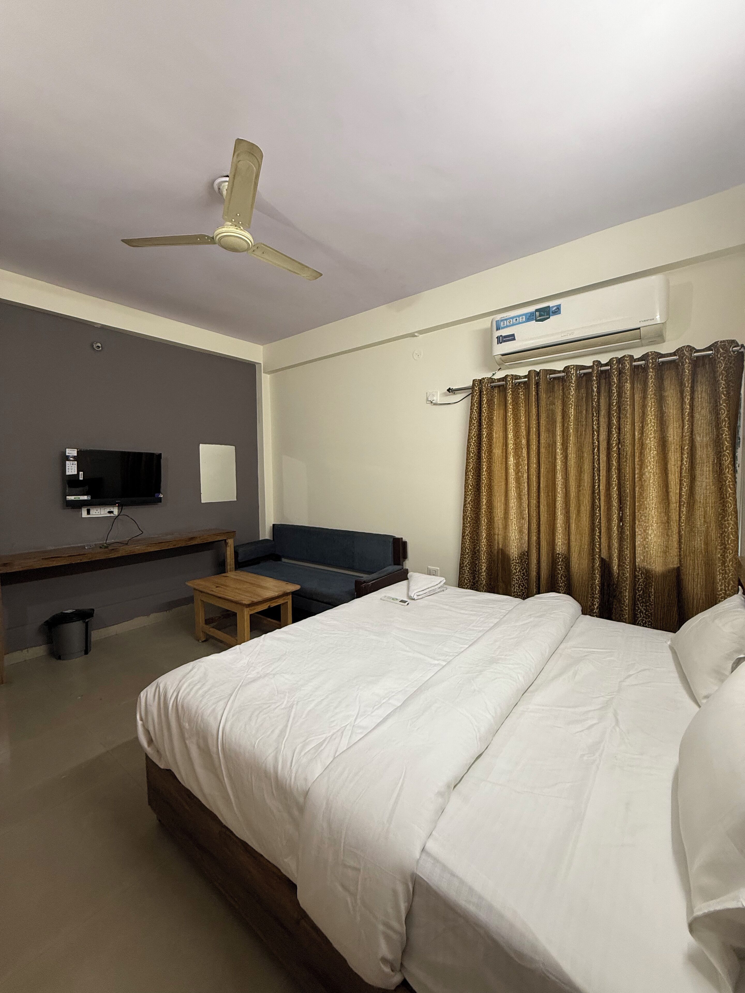 A hotel room with a bed, a desk, a chair, a ceiling fan, an air conditioner, and a television.