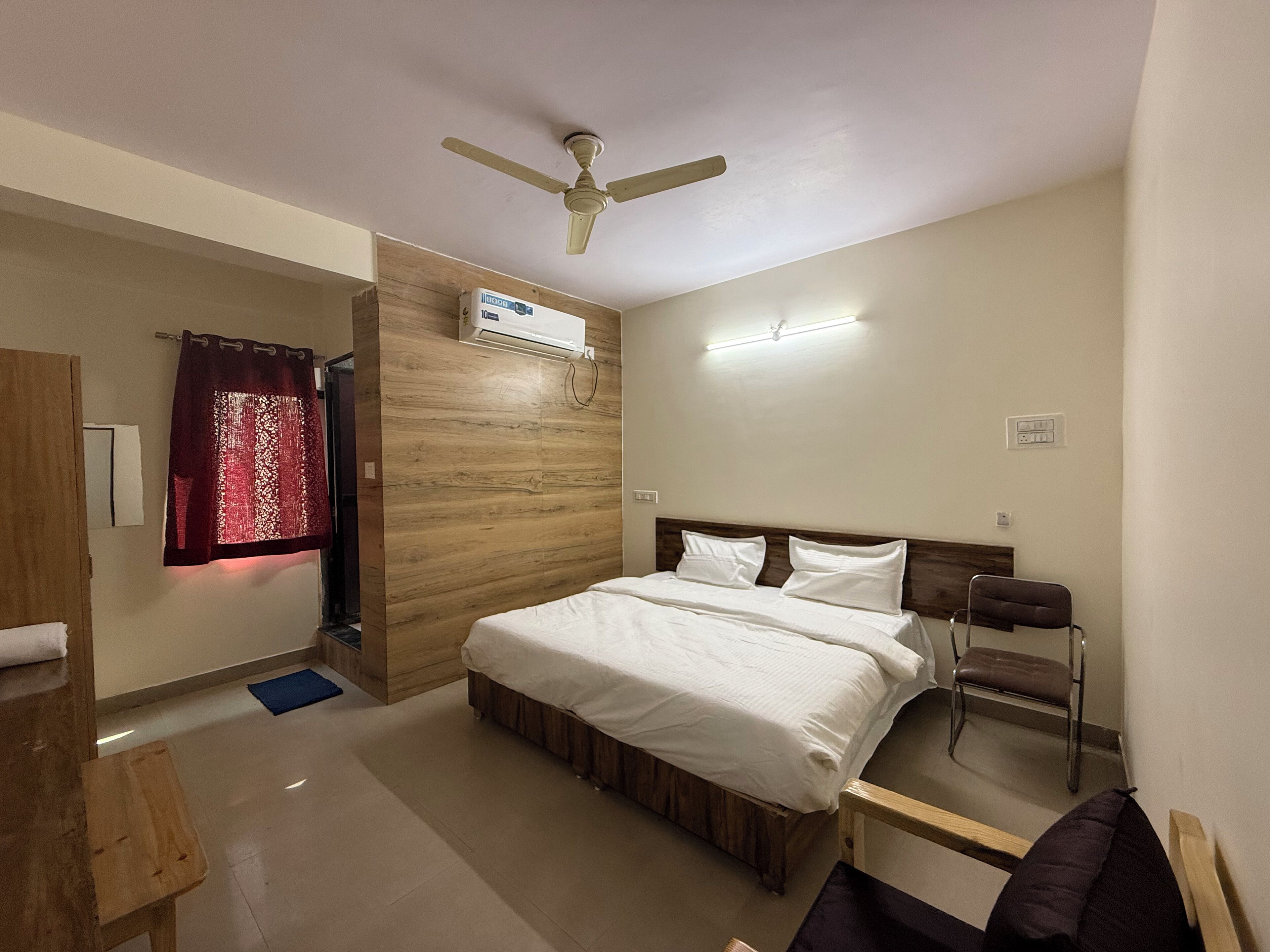 A bedroom with a bed, a ceiling fan, an air conditioning unit, a bedside table, a chair, and a wooden wardrobe.