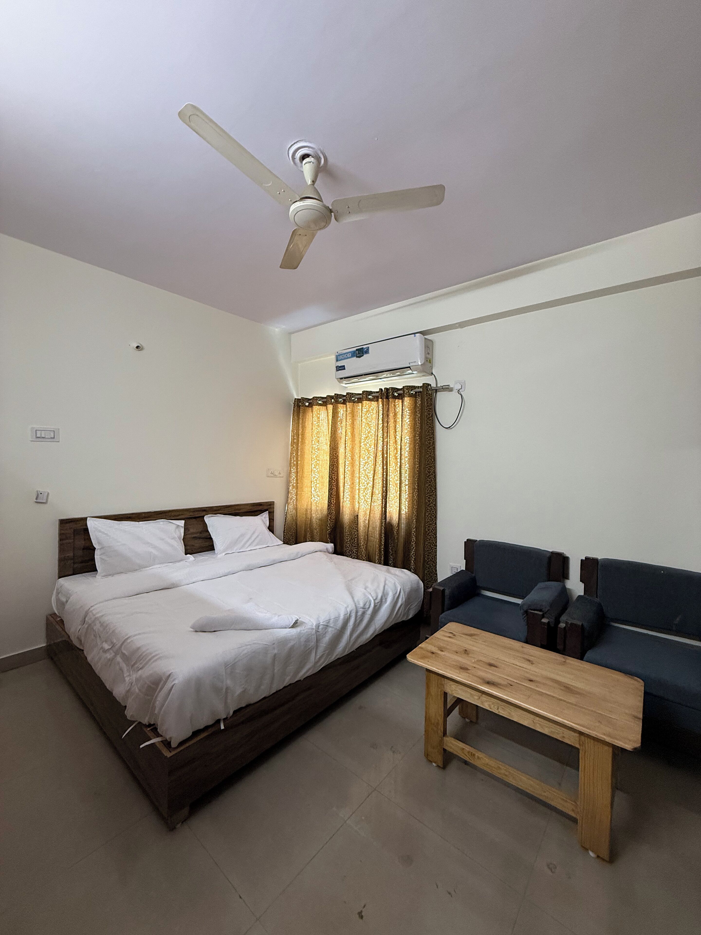 A bedroom with a bed, a ceiling fan, an air conditioning unit, a sofa, and a wooden table.