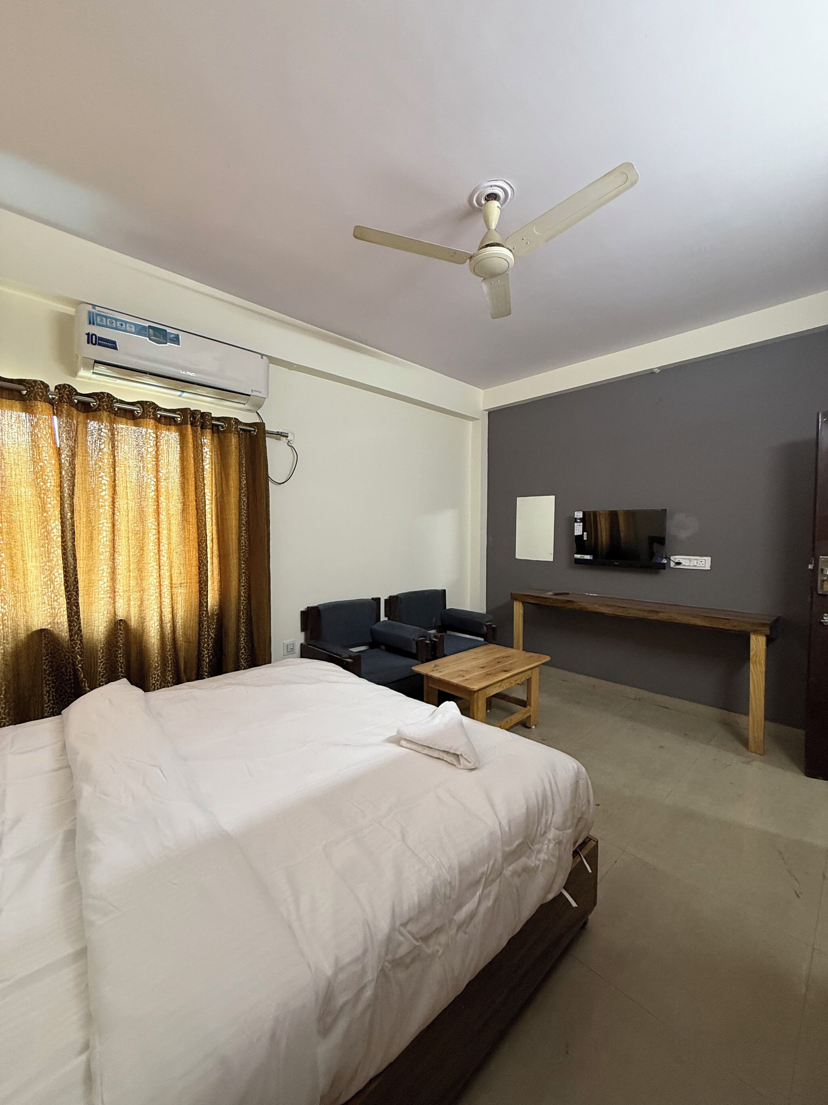A hotel room with a bed, a desk, a chair, a ceiling fan, and an air conditioning unit.