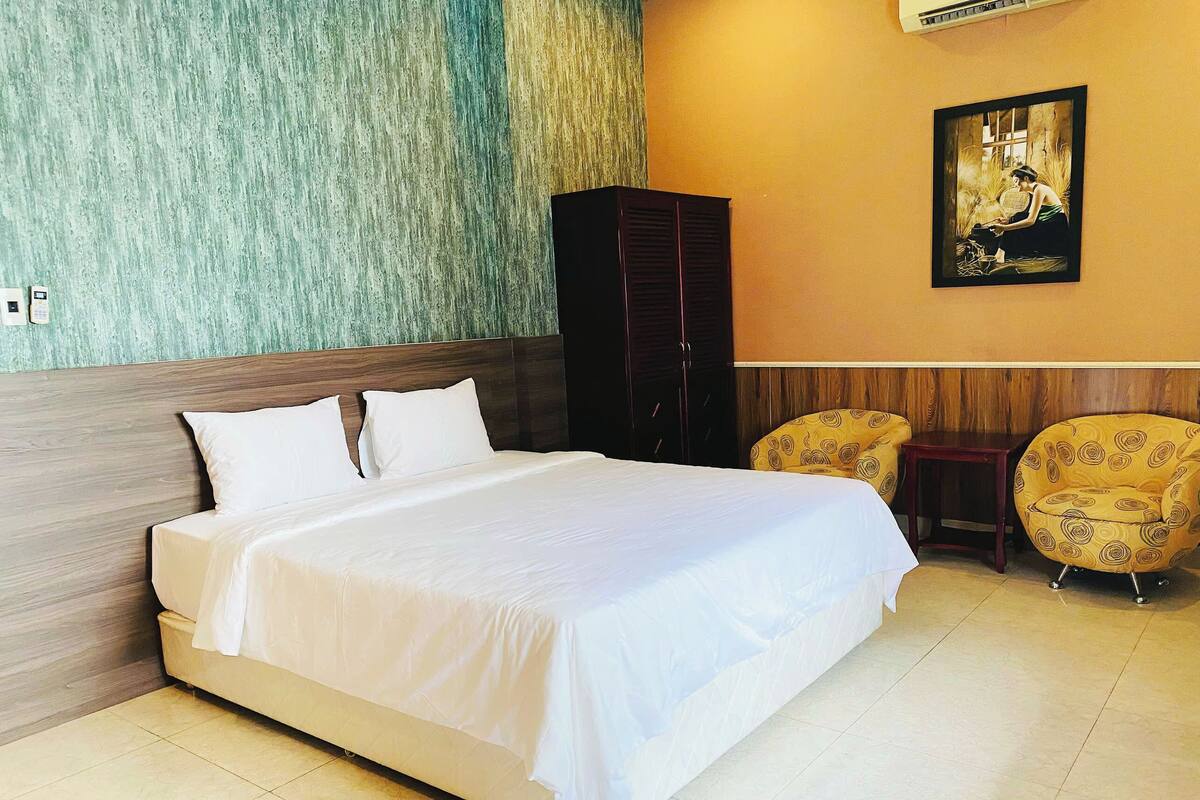 A hotel room with a large bed, two armchairs, a wooden wardrobe, and a framed picture on the wall.