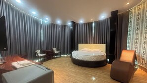 Ein modernes Hotelzimmer mit einem runden Bett, Holzboden, Schreibtisch mit Stuhl und einem großen Fenster mit Vorhängen.
