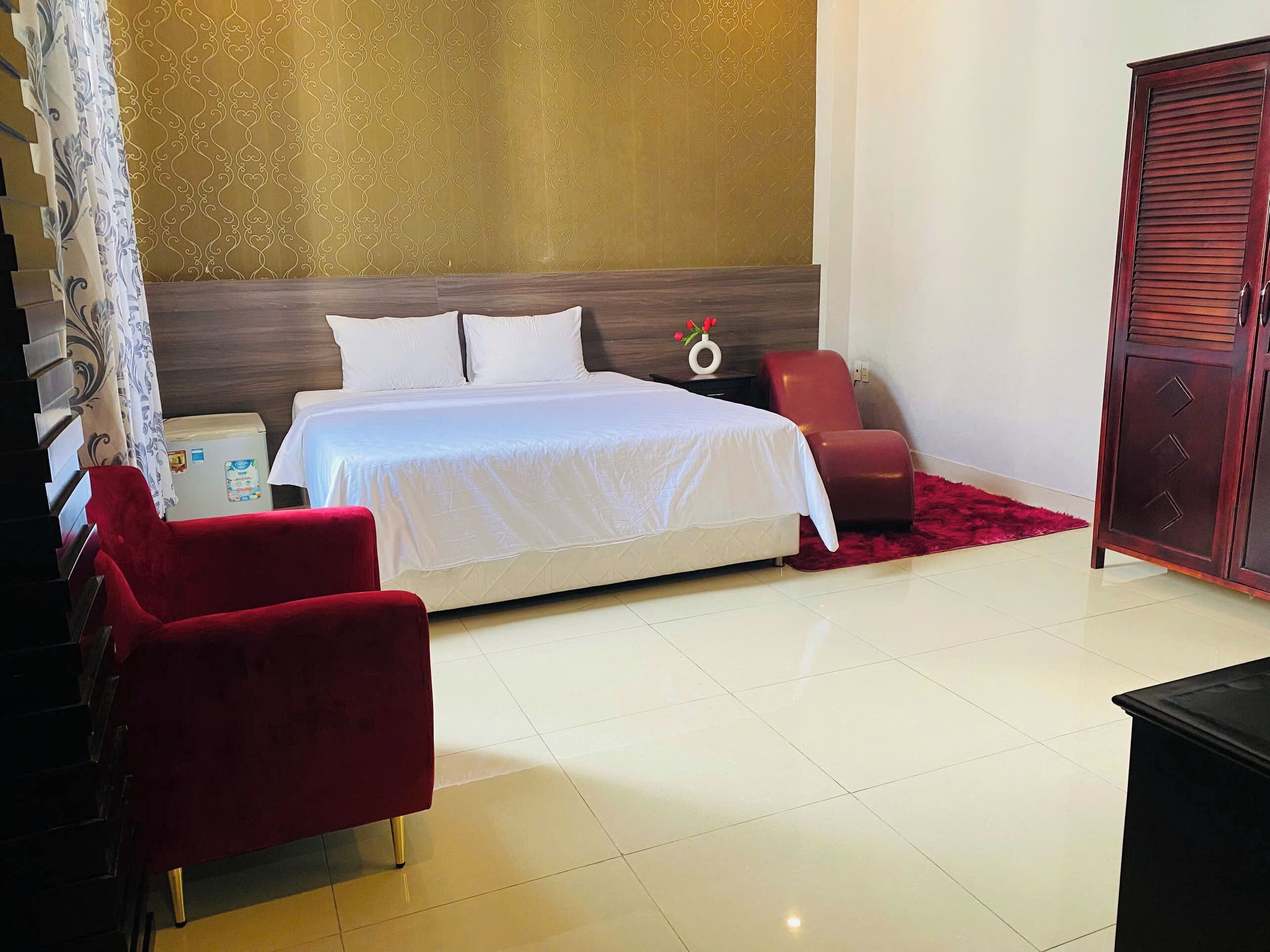 Una habitación de hotel con una cama, un sillón rojo, un armario de madera y una mesita de noche con un jarrón de flores.