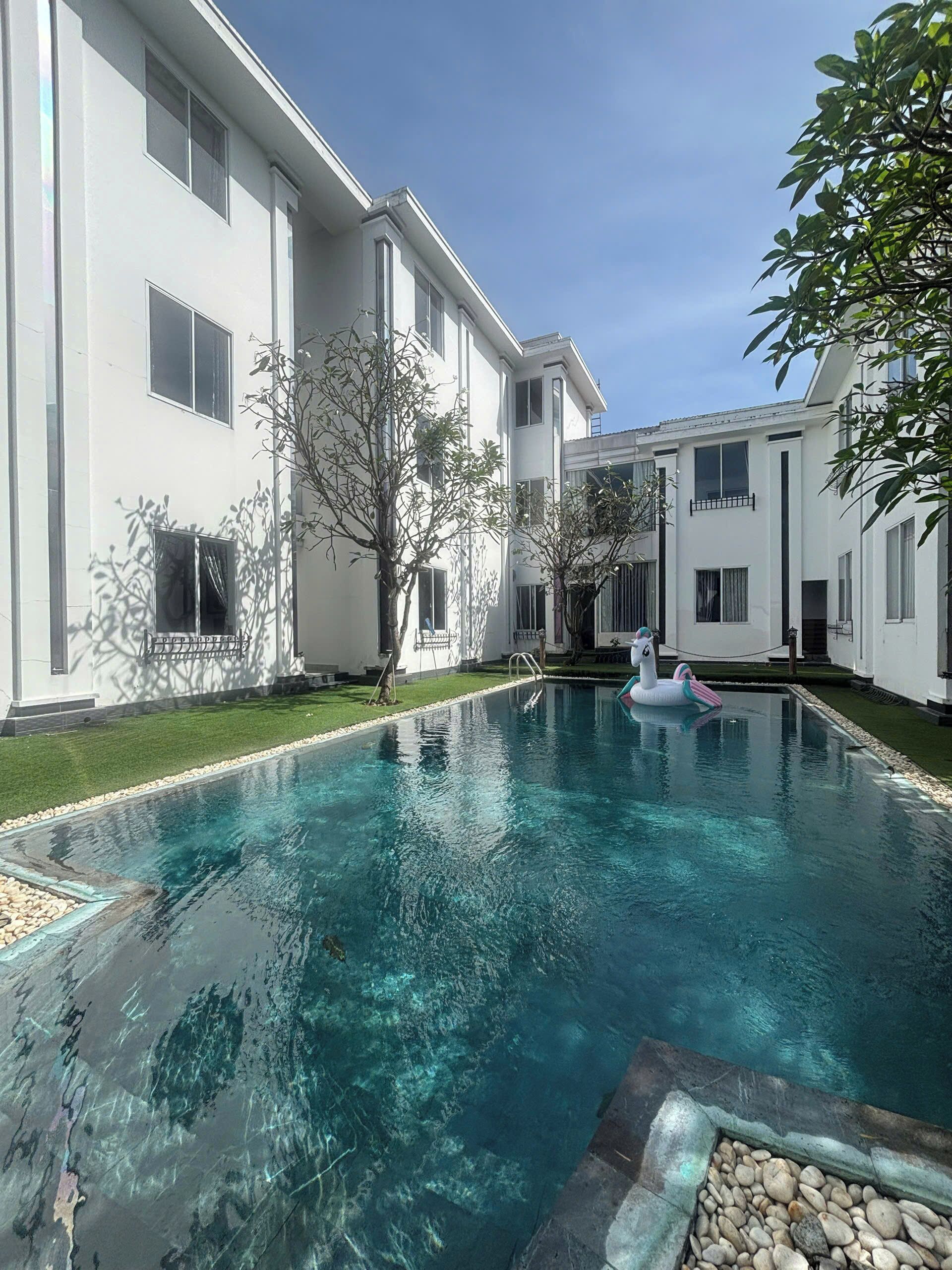 A swimming pool with clear water, surrounded by a white building with multiple windows and a small tree.