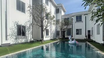 A swimming pool with clear water, surrounded by a white building with multiple windows and a small tree.