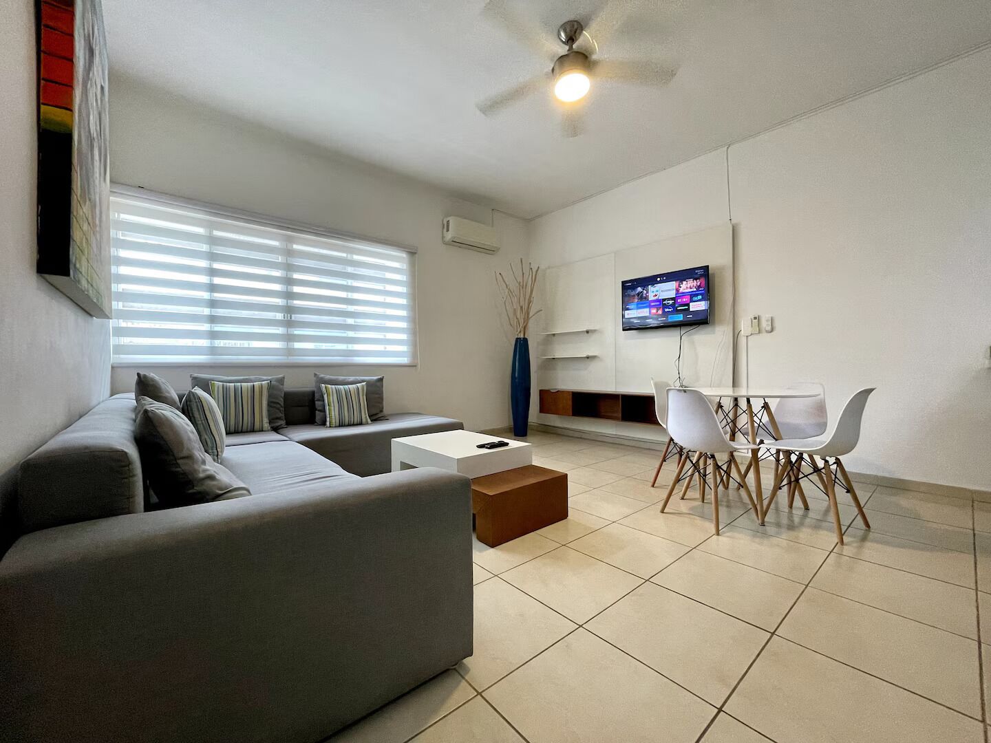 Ein modernes Wohnzimmer mit grauem Sofa, einem Couchtisch, einem Esstisch mit Stühlen, einem an der Wand befestigten Fernseher und einem Deckenventilator.