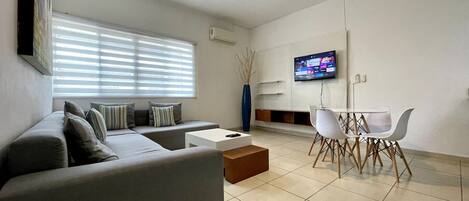 Ein modernes Wohnzimmer mit grauem Sofa, einem Couchtisch, einem Esstisch mit Stühlen, einem an der Wand befestigten Fernseher und einem Deckenventilator.