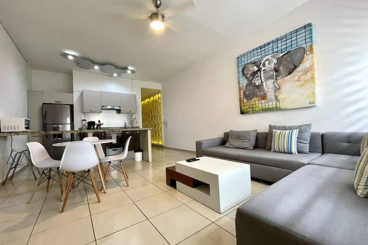 A modern living room with a grey sofa, a coffee table, and a dining area with chairs and a kitchen in the background.