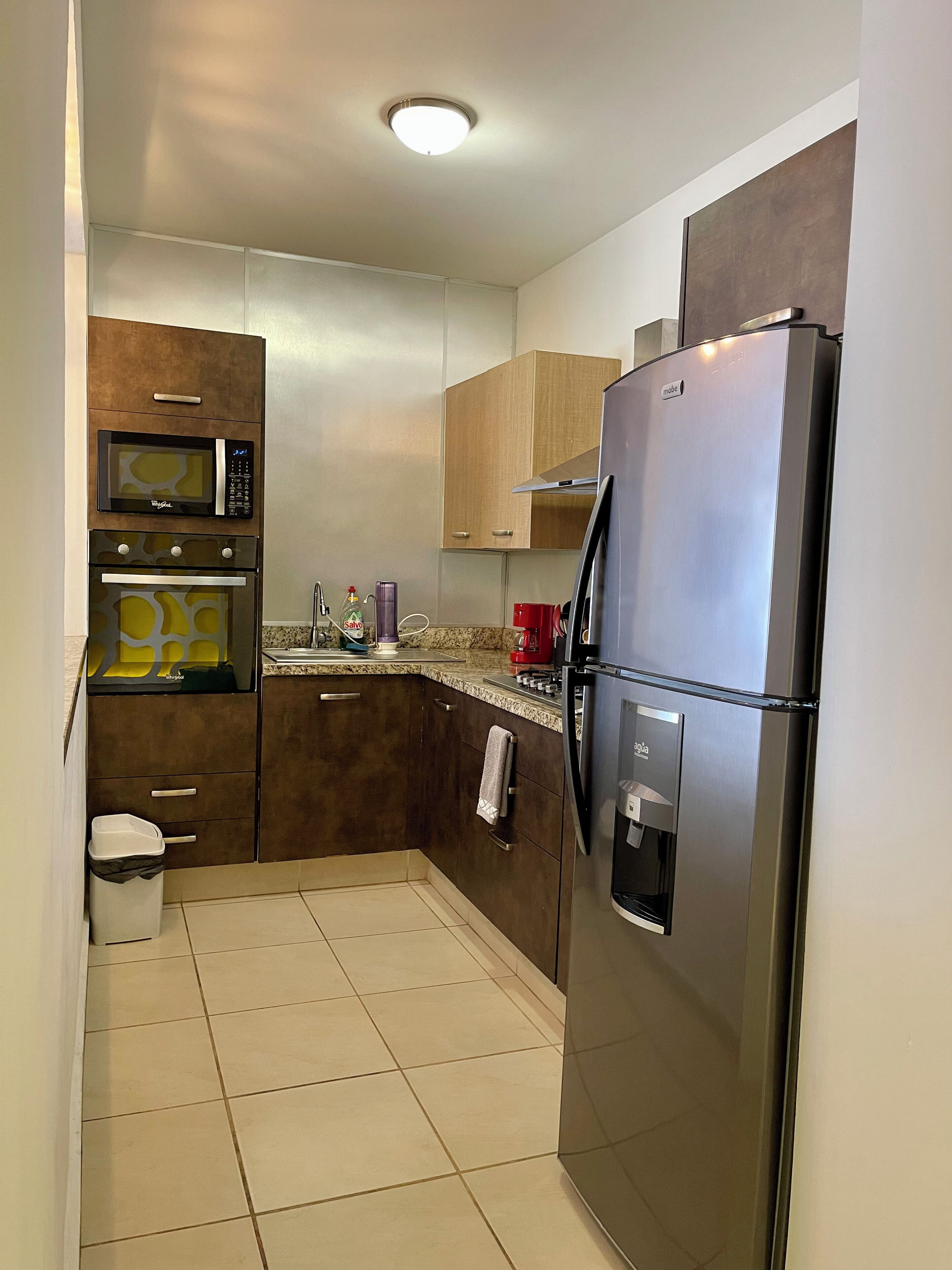 A compact kitchen with a stainless steel refrigerator, a microwave, and a built-in oven. There is a trash bin and a towel hanging on the wall.