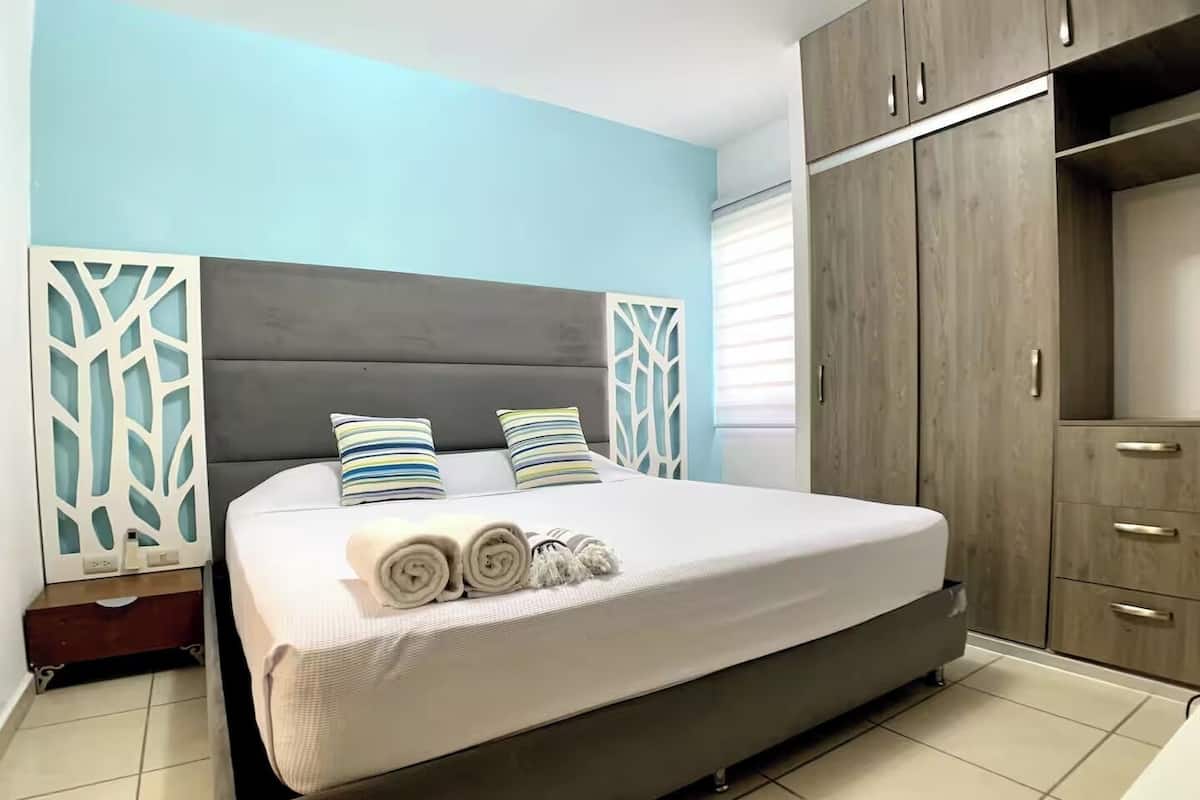 A modern bedroom with a large bed, two pillows, a nightstand, a wardrobe, and a window with blinds.