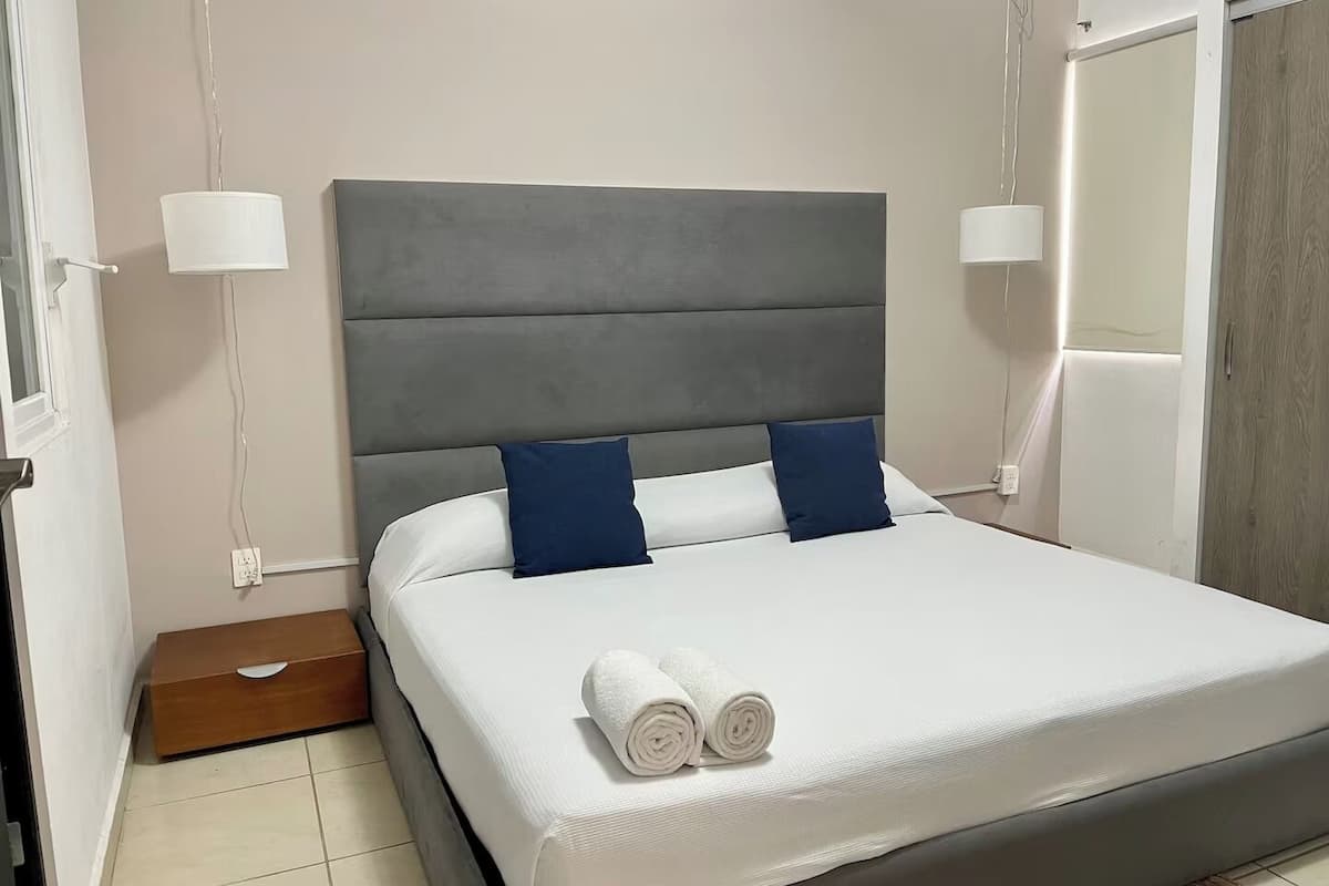 A neatly made bed with white linens and two dark blue pillows, a grey upholstered headboard, and bedside lamps on either side.