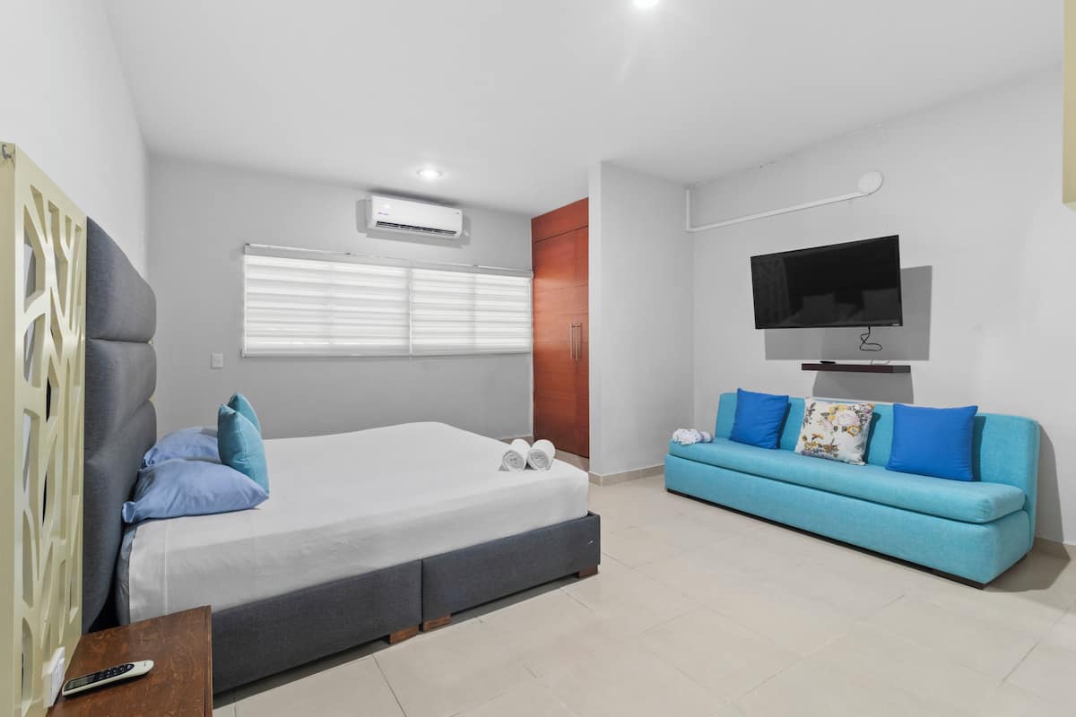 A modern bedroom with a bed, a TV, a sofa, and a window with blinds.
