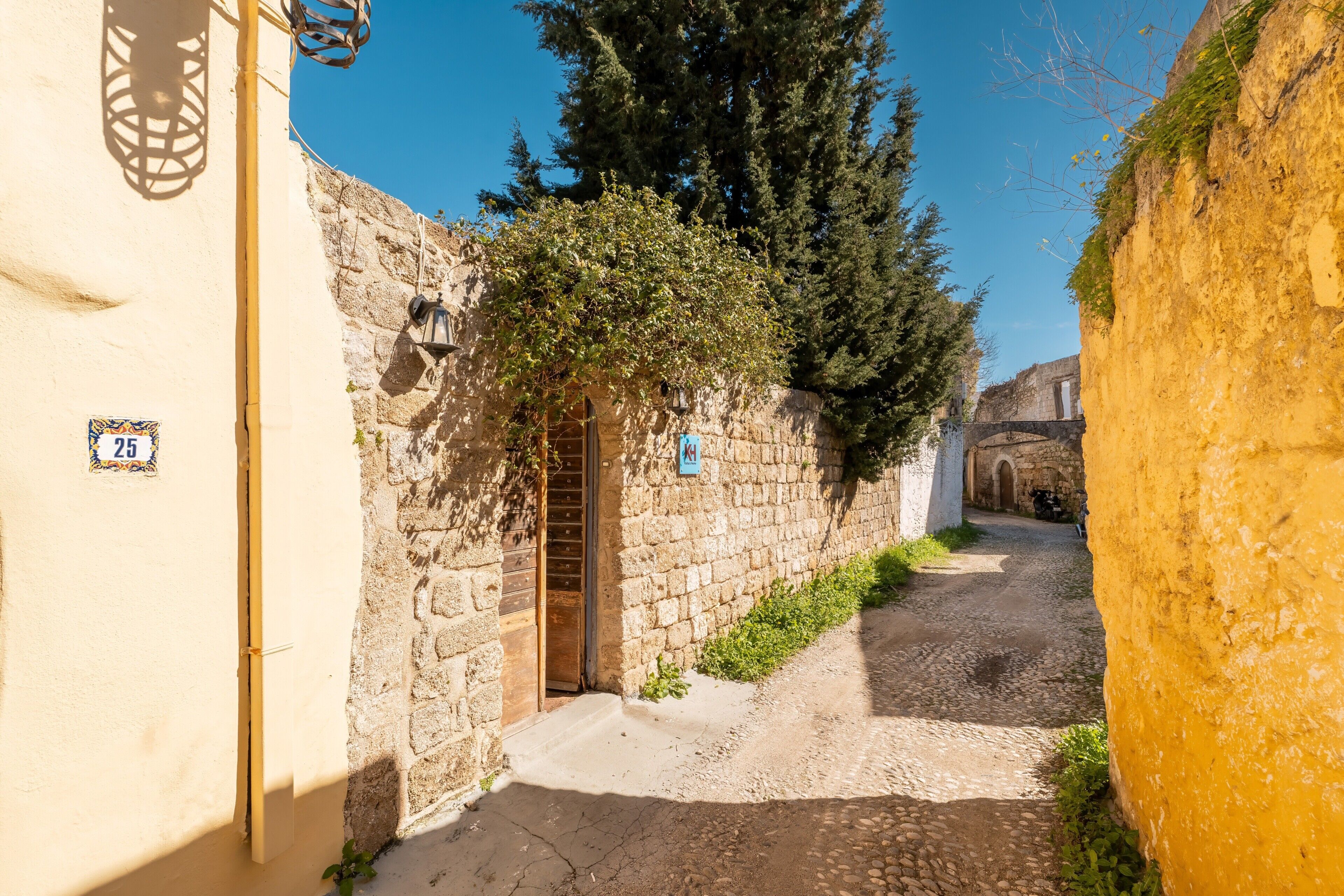 Une ruelle étroite pavée de pierres, bordée de murs en pierre, avec une porte en bois et un panneau indiquant une zone piétonne.