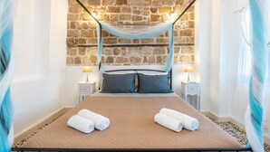 A bed with a canopy, two bedside lamps, a stone wall, and two rolled towels on the bed. - Holiday Home 'Katia's Home First Floor' with Shared Garden, Wi-Fi and Air Conditioning (Rhodes)