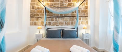 A bed with a canopy, two bedside lamps, a stone wall, and two rolled towels on the bed.