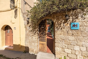 Un edificio in pietra con una porta in legno e un arco, una targa con su scritto 'Katia's Home' e il numero '27' sulla parete.