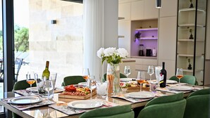Una mesa de comedor dispuesta con platos, vasos, comida y bebidas variadas, sillas verdes y un florero con flores.