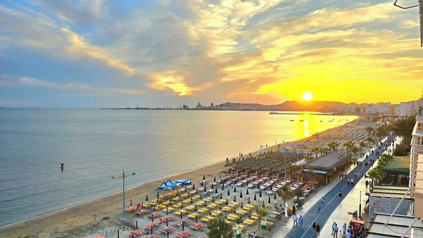 Una playa con hileras de sombrillas y tumbonas, un paseo marítimo y una puesta de sol sobre el agua.