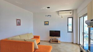 Una sala de estar moderna con un sofá grande color naranja, un televisor de pantalla plana sobre un soporte de madera y vistas a la ciudad a través de un ventanal.