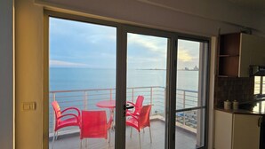 Un balcón con sillas rojas y una mesa, con vista a una masa de agua y un horizonte urbano lejano.