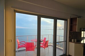Un balcón con sillas rojas y una mesa, con vista a una masa de agua y un horizonte urbano distante.