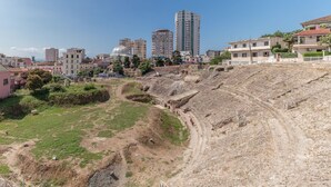 Um grande anfiteatro ao ar livre com palco de terra e área de assentos, rodeado por edifícios modernos e vegetação.
